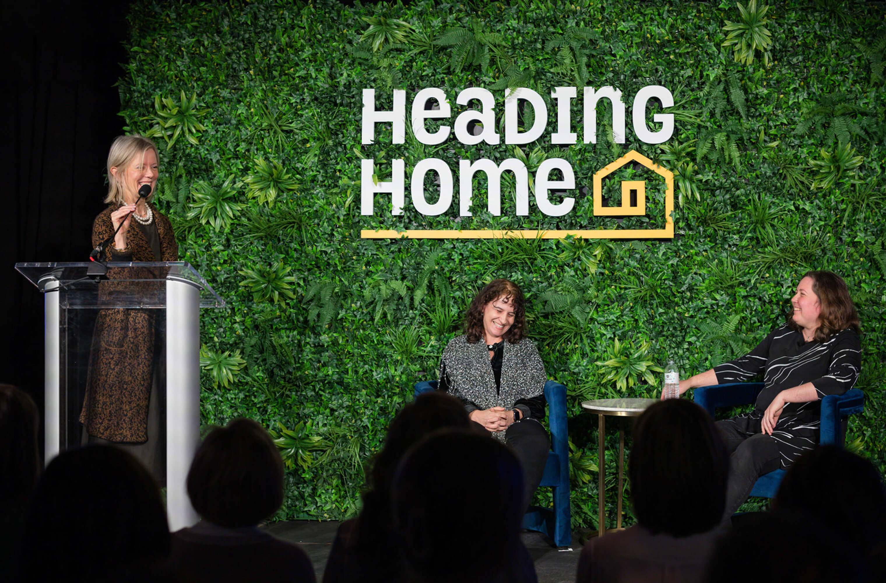 Speakers on stage in front of a Heading Home greenery wall at a fundraising event