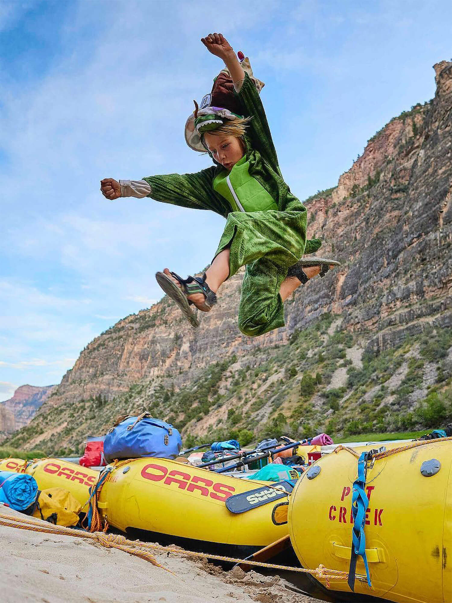 A child in a dinosaur costume leaping beside OARS rafts on a canyon beach